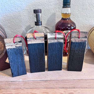 May include: Four rectangular wooden coasters with a dark blue finish and red string loops. The coasters are made from reclaimed wood and are displayed on a wooden surface, with whiskey bottles and a barrel in the background.