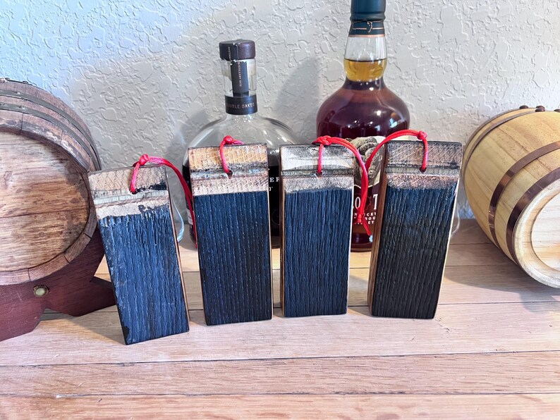 May include: Four rectangular wooden coasters with a dark blue finish and red string loops. The coasters are made from reclaimed wood and are displayed on a wooden surface, with whiskey bottles and a barrel in the background.