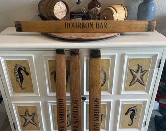 May include: A white cabinet with seahorse and starfish designs, topped with a wooden bourbon bar sign and small barrels. Three wooden bourbon bar staves stand in front. Bottles of liquor and a lamp are also visible.