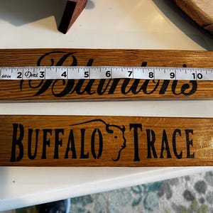 Puede incluir: Dos letreros de madera con letras oscuras. El letrero superior dice "Sullivan's" y mide aproximadamente 30 cm de largo. El letrero inferior dice "Buffalo Trace" con un gr&aacute;fico de cabeza de b&uacute;falo. Ambos letreros est&aacute;n hechos de madera te&ntilde;ida.
