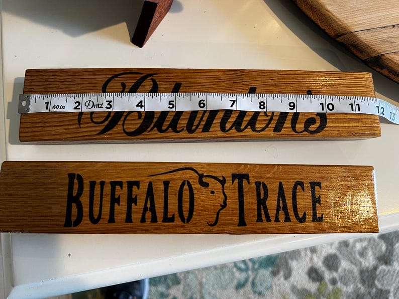Puede incluir: Dos letreros de madera con letras oscuras. El letrero superior dice "Sullivan's" y mide aproximadamente 30 cm de largo. El letrero inferior dice "Buffalo Trace" con un gr&aacute;fico de cabeza de b&uacute;falo. Ambos letreros est&aacute;n hechos de madera te&ntilde;ida.