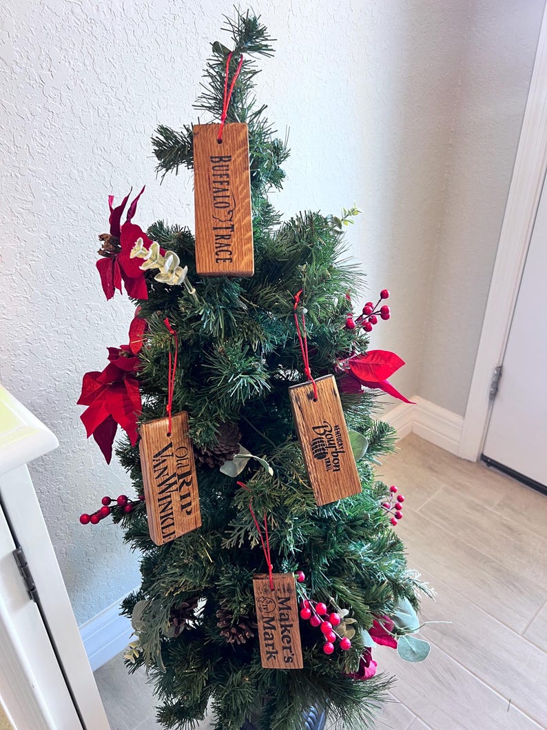 May include: A small artificial Christmas tree decorated with wooden ornaments. The ornaments are rectangular and feature the names "Buffalo Trace", "Old Rip Van Winkle", "Kentucky Bourbon", and "Maker's Mark". Red berries and poinsettias add color.