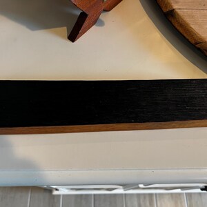 Puede incluir: Una pieza rectangular de madera con la parte superior negra y la inferior de color madera natural. La veta de la madera es visible en la superficie negra. El art&iacute;culo parece ser una pieza decorativa o funcional.