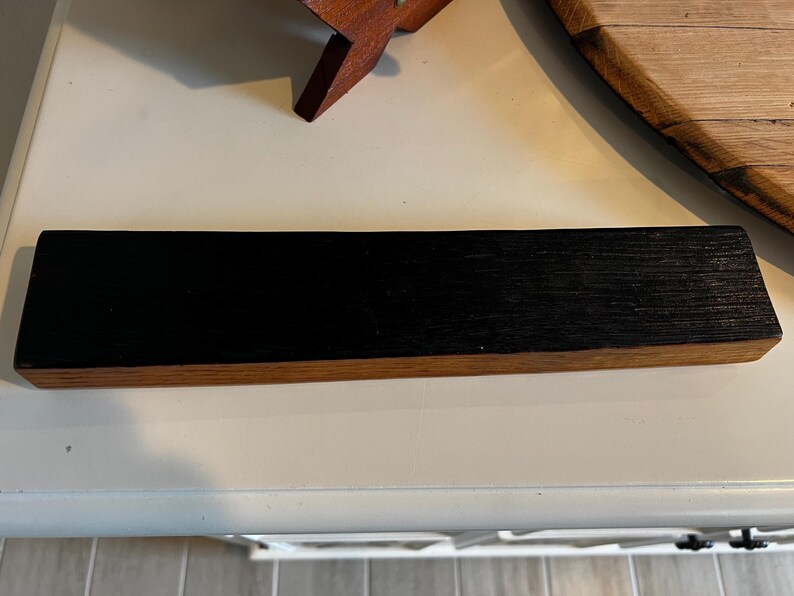 Puede incluir: Una pieza rectangular de madera con la parte superior negra y la inferior de color madera natural. La veta de la madera es visible en la superficie negra. El art&iacute;culo parece ser una pieza decorativa o funcional.