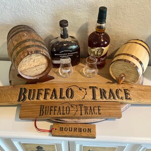 Puede incluir: Una exhibici&oacute;n de art&iacute;culos relacionados con el bourbon, que incluye dos peque&ntilde;os barriles de madera, dos botellas de bourbon, dos vasos de chupito y letreros de madera que dicen "Buffalo Trace" y "Bourbon". Los art&iacute;culos est&aacute;n dispuestos sobre una superficie blanca.