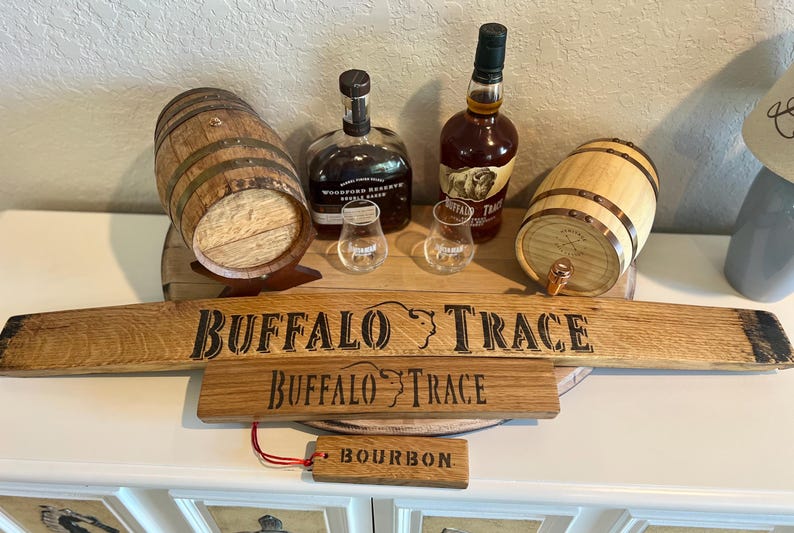 Puede incluir: Una exhibici&oacute;n de art&iacute;culos relacionados con el bourbon, que incluye dos peque&ntilde;os barriles de madera, dos botellas de bourbon, dos vasos de chupito y letreros de madera que dicen "Buffalo Trace" y "Bourbon". Los art&iacute;culos est&aacute;n dispuestos sobre una superficie blanca.