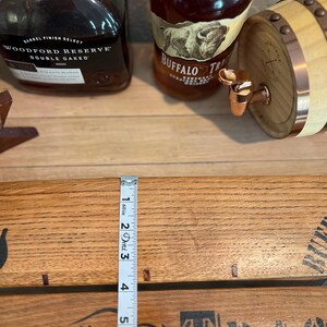 May include: A wooden barrel with a copper spigot, two bottles of whiskey, and a wooden plank with the text "BUFFALO TRACE". A measuring tape shows a measurement of 6 inches.