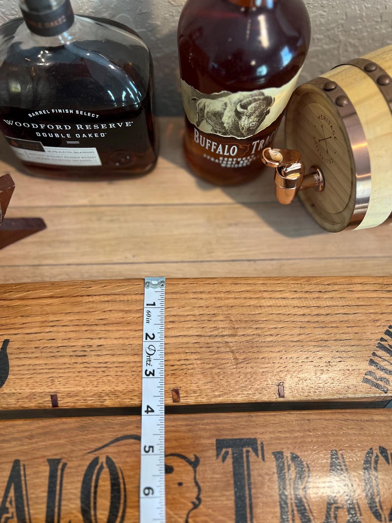May include: A wooden barrel with a copper spigot, two bottles of whiskey, and a wooden plank with the text "BUFFALO TRACE". A measuring tape shows a measurement of 6 inches.
