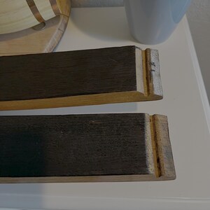 Op de afbeelding: Twee rechthoekige houten stukken met donkerbruine bovenkanten en lichtbruine zijkanten. De uiteinden hebben een gekerfd ontwerp. De houtnerf is zichtbaar. De stukken lijken deel uit te maken van een grotere houten structuur.