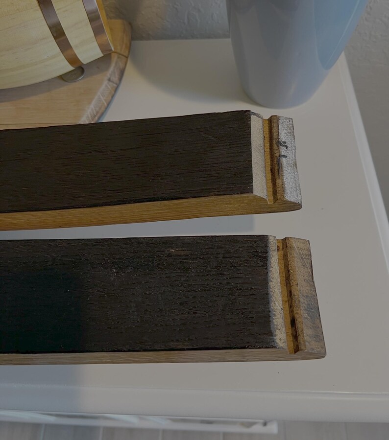 Op de afbeelding: Twee rechthoekige houten stukken met donkerbruine bovenkanten en lichtbruine zijkanten. De uiteinden hebben een gekerfd ontwerp. De houtnerf is zichtbaar. De stukken lijken deel uit te maken van een grotere houten structuur.