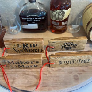 May include: Wooden bourbon barrel stave tags with the names "Old Rip Van Winkle", "Kentucky Bourbon Trail", "Maker's Mark", and "Buffalo Trace" printed on them. The tags are attached with red string. Bourbon bottles and a small barrel are in the background.