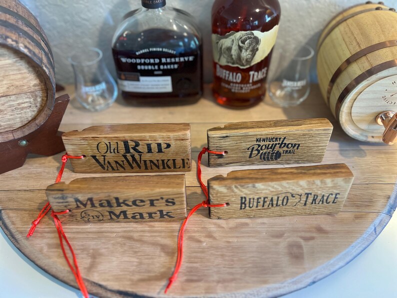 May include: Wooden bourbon barrel stave tags with the names "Old Rip Van Winkle", "Kentucky Bourbon Trail", "Maker's Mark", and "Buffalo Trace" printed on them. The tags are attached with red string. Bourbon bottles and a small barrel are in the background.