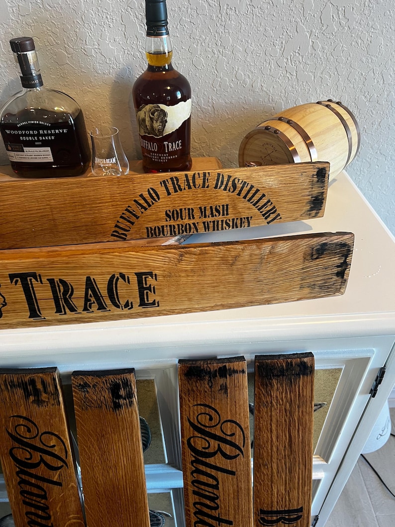 May include: Wooden bourbon barrel stave signs with the text "Buffalo Trace Distillery" and "Trace" in black lettering. Two bottles of bourbon and a small barrel are also visible. The signs are made from reclaimed wood.