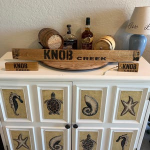 May include: A white cabinet with six framed panels featuring sea life designs. The cabinet is topped with a wooden sign that reads "KNOB CREEK", along with two small barrels and two bottles of liquor. A lamp is visible in the background.