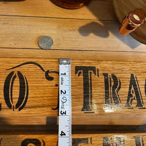 May include: A wooden sign with the words "Buffalo Trace" in black lettering. A measuring tape shows a measurement of approximately 4 inches. A bottle of bourbon and a coin are also visible.