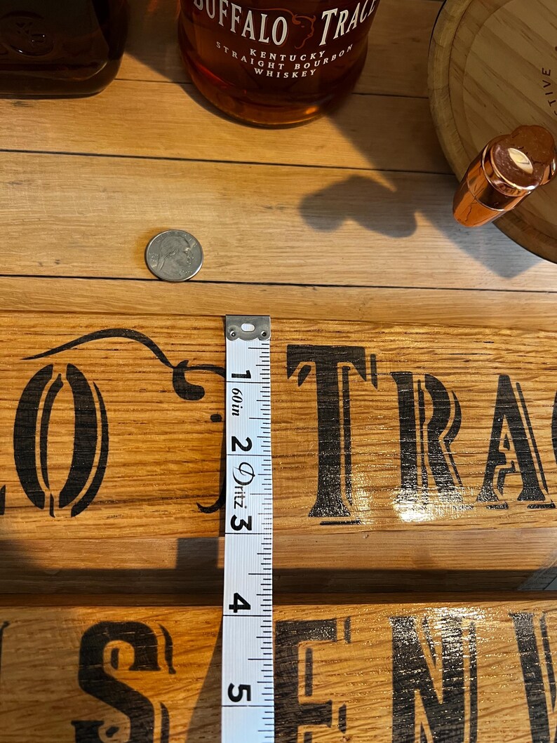 May include: A wooden sign with the words "Buffalo Trace" in black lettering. A measuring tape shows a measurement of approximately 4 inches. A bottle of bourbon and a coin are also visible.