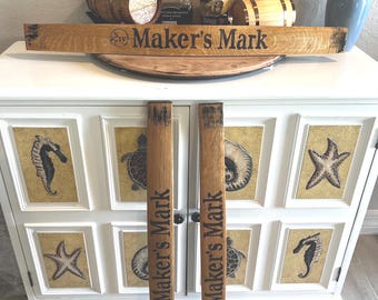 May include: A white cabinet with framed sea life art, topped with a wooden sign that reads "Maker's Mark". Two wooden barrel staves with "Maker's Mark" printed on them are in front of the cabinet. Bottles and small barrels are on top.