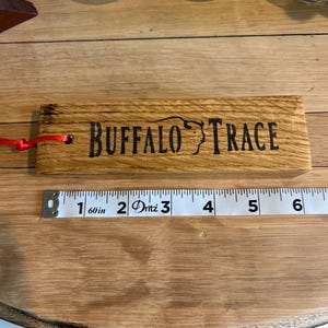 May include: A wooden Buffalo Trace branded item with a red string attached. The text is etched in black. A measuring tape shows the item is approximately 6 inches long. Whiskey glasses are in the background.