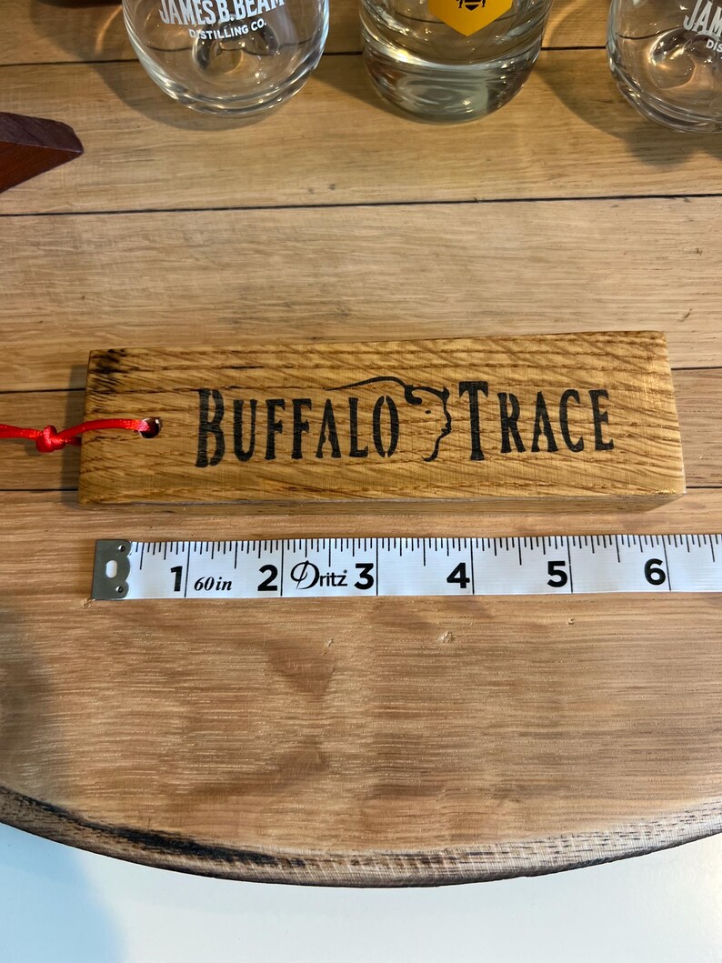 May include: A wooden Buffalo Trace branded item with a red string attached. The text is etched in black. A measuring tape shows the item is approximately 6 inches long. Whiskey glasses are in the background.