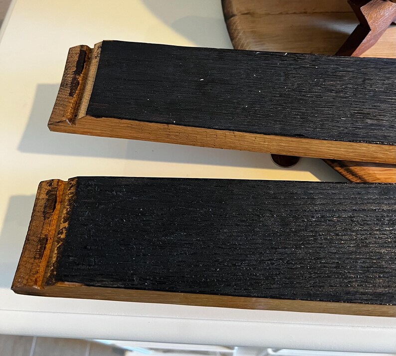 Op de afbeelding: Twee rechthoekige houten serveerplanken. De planken hebben een donker, getextureerd oppervlak dat wordt omlijst door lichter gekleurd hout. De uiteinden van de planken hebben een decoratief, gekerfd ontwerp. De planken zijn waarschijnlijk bedoeld om voedsel te serveren.