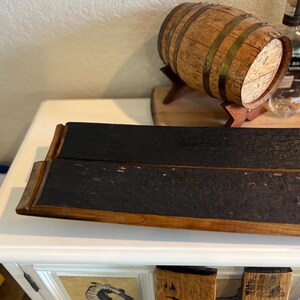 May include: A wooden barrel with metal bands and a small wooden stand. A dark, rectangular serving tray with a charred appearance is in the foreground. The word "Bla" is visible on a wooden piece.