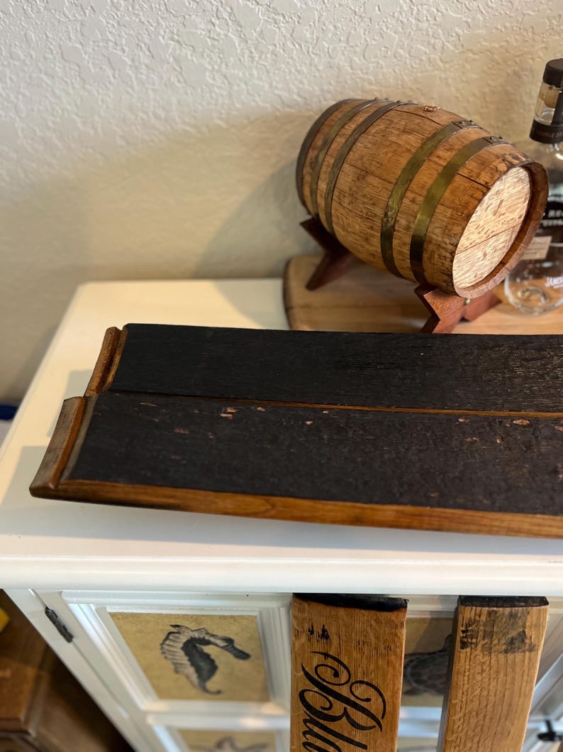 May include: A wooden barrel with metal bands and a small wooden stand. A dark, rectangular serving tray with a charred appearance is in the foreground. The word "Bla" is visible on a wooden piece.
