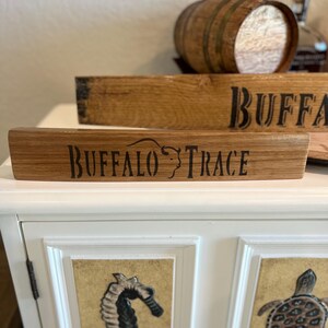 Puede incluir: Letrero de madera con el texto "BUFFALO TRACE" en letras negras. El letrero est&aacute; hecho de madera de color marr&oacute;n claro y tiene un aspecto r&uacute;stico. Un peque&ntilde;o barril de madera est&aacute; en el fondo. El letrero probablemente est&eacute; relacionado con el bourbon o el whisky.
