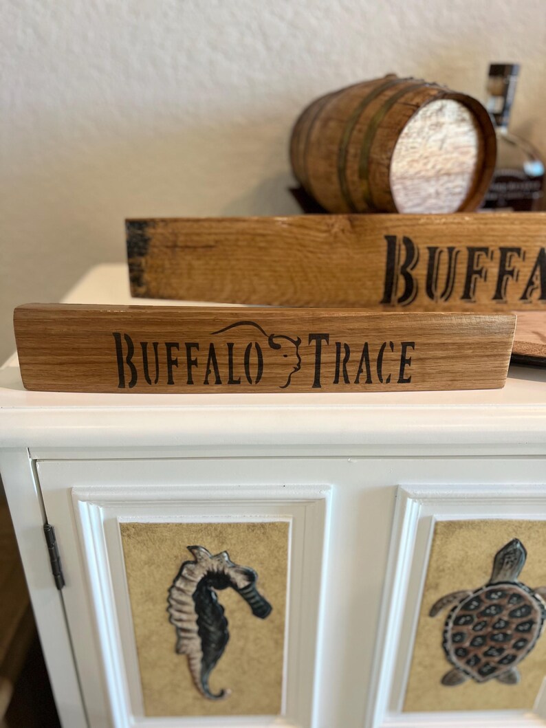 Puede incluir: Letrero de madera con el texto "BUFFALO TRACE" en letras negras. El letrero est&aacute; hecho de madera de color marr&oacute;n claro y tiene un aspecto r&uacute;stico. Un peque&ntilde;o barril de madera est&aacute; en el fondo. El letrero probablemente est&eacute; relacionado con el bourbon o el whisky.