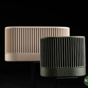 May include: Two cylindrical decorative objects with vertical ribbed detailing. One is a light beige color, and the other is a dark olive green. Both have a solid base and are set against a black background. The objects appear to be made of a matte material.