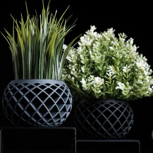 May include: Two decorative planters with artificial plants. The planters are dark gray, spherical, and feature a diamond-patterned design. One planter holds tall, green grass-like plants, and the other holds a bush with small white flowers.