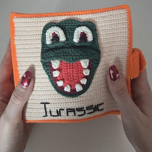 May include: Crocheted book with a dinosaur face on the cover. The dinosaur is green with white teeth and a red mouth. The word "JURASSIC" is stitched below the dinosaur. The book has an orange border and a small orange loop on the side.