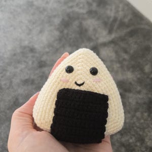 May include: A crocheted onigiri plushie in the shape of a rice ball. The plushie is off-white with a black seaweed wrap, black button eyes, and pink cheeks. The onigiri has a smiling face.