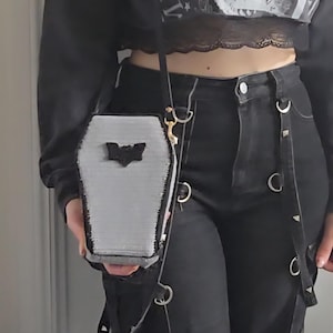 Può includere: Una borsa a tracolla a forma di bara grigia e nera, con un emblema di pipistrello nero. La borsa ha una superficie strutturata e una tracolla nera con finiture color oro. La persona indossa jeans neri con anelli metallici color argento.