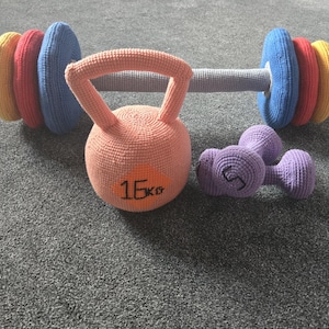 May include: A collection of colorful, knitted exercise equipment, including a barbell with yellow, red, and blue weights, a peach-colored kettlebell marked "16KG", and a pair of purple dumbbells. The items are arranged on a gray surface.