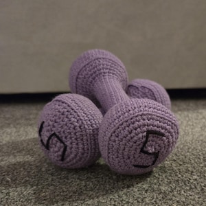 May include: A set of three crochet dumbbells in a soft lavender color. Each dumbbell has a black number 5 stitched on the side. The dumbbells are made of a textured yarn and are resting on a gray surface.