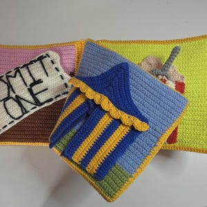 Puede incluir: Un libro sensorial de ganchillo hecho a mano con páginas de temática de feria. Una página muestra un fondo marrón y rosa con un parche de ticket blanco con el texto ADMIT ONE. Otra página muestra una carpa azul y amarilla a rayas.