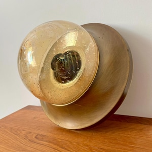 Może przedstawiać: Wintageowy kinkiet ścienny z kulistym, nakrapianym szklanym kloszem w ciepłym bursztynowym odcieniu, osadzony na okrągłej, mosiężnej metalowej podstawie. Projekt ma estetykę modernizmu z połowy wieku, z naciskiem na geometryczne kształty i ciepłe odcienie.