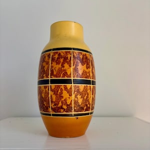 May include: A vintage ceramic vase with a cylindrical shape. The vase features a yellow top and base, with a central section adorned with a brown and orange patterned design, framed by black horizontal lines. The vase is a decorative item.