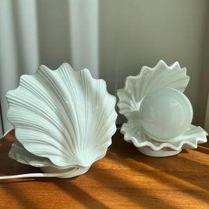 Könnte beinhalten: Zwei weiße, muschelförmige Keramiklampen. Eine Lampe ist geschlossen, die andere offen und zeigt eine Glühbirne. Die Lampen stehen auf einer Holzoberfläche, mit einem weißen Kabel. Die Lampen haben ein strukturiertes, geriffeltes Design.