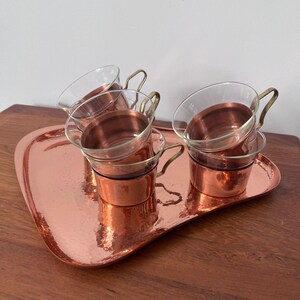 Könnte beinhalten: Ein Set aus vier Kupfer-Glas-Tassen mit Messinggriffen, das auf einem passenden Kupfertablett steht. Das Tablett hat eine gehämmerten Textur und eine einzigartige, geschwungene Form. Die Tassen sind aus klarem Glas mit Kupferböden.