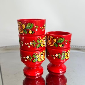 Könnte beinhalten: Vier rote, handbemalte Eierbecher aus Holz mit gelben, grünen und weißen Blumenmustern. Die Eierbecher sind gestapelt und stehen auf einer reflektierenden Oberfläche.