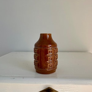 Könnte beinhalten: Eine braune Keramikvase mit glänzendem Finish. Die Vase hat eine Reihe von quadratischen, geprägten Designs um ihren Körper. Sie hat einen abgerundeten Boden und einen leicht verjüngten Hals, geeignet für die Inneneinrichtung.