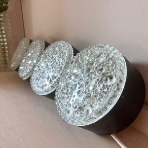 Könnte beinhalten: Eine Reihe von vier Vintage-Wandleuchten. Jede Leuchte hat einen schwarzen zylindrischen Sockel und einen transparenten, strukturierten Glasschirm. Die Schirme sind mit kleinen, blasenartigen Vorsprüngen bedeckt. Ein weißes Elektrokabel erstreckt sich von den Leuchten.