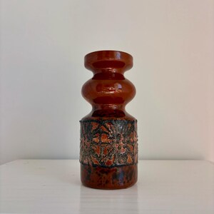 May include: A brown ceramic vase with a textured, patterned design. The vase has a glossy finish and a bulbous shape with a narrow neck. The textured section features a mix of orange, black, and brown colors.