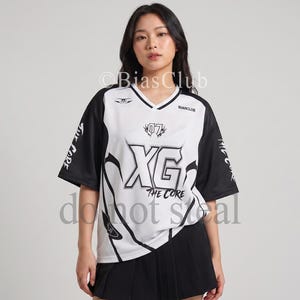 Può includere: Una maglia bianca e nera a maniche corte con scollo a V. La maglia presenta il testo "XG THE CORE" e il numero "07" in grassetto. Le maniche sono nere con testo bianco. La modella indossa una gonna a pieghe nera.