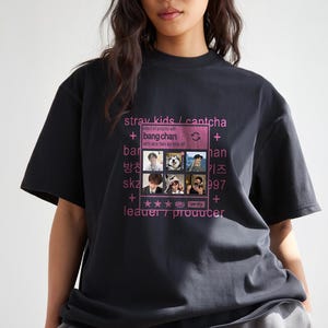 May include: Black t-shirt featuring a pink graphic with the text "stray kids / cantcha" and images of band members. The design includes the words "bang chan", "leader / producer", and other text.