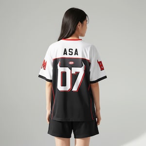 Puede incluir: Camiseta deportiva blanca y negra con el texto "ASA" y el número "07" en rojo. La camiseta tiene ribetes rojos y mangas negras. La modelo lleva pantalones cortos negros.