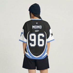 Puede incluir: Camiseta negra estilo jersey con detalles en blanco y azul, con el nombre "MOMO" y el n&uacute;mero "96" en blanco. La camiseta tiene mangas cortas y cuello redondo. Un gorro negro y pantalones cortos negros completan el look.