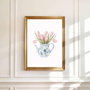 以下が含まれることがあります： ピンクのチューリップが入った青と白の花柄のティーポットが描かれた、額入りの水彩画プリント。アートワークは白い背景に配置され、金色のフレームに額装されており、クラシックでエレガントな美しさを生み出しています。