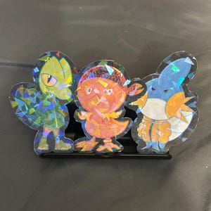 May include: Three holographic stickers of cartoon characters. The stickers have a colourful, iridescent finish. The characters are standing on a black stand. The stickers are in the shape of the characters.